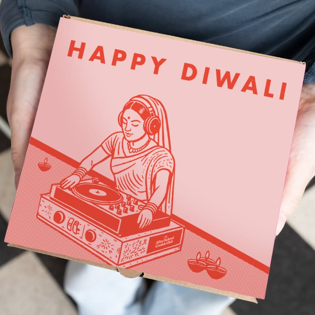 A person holding a square pink and red printed box with the text 'HAPPY DIwali' and an illustration of a woman operating a turntable. The person is wearing a dark blue-gray long-sleeve top and white pants, standing on a tiled floor.