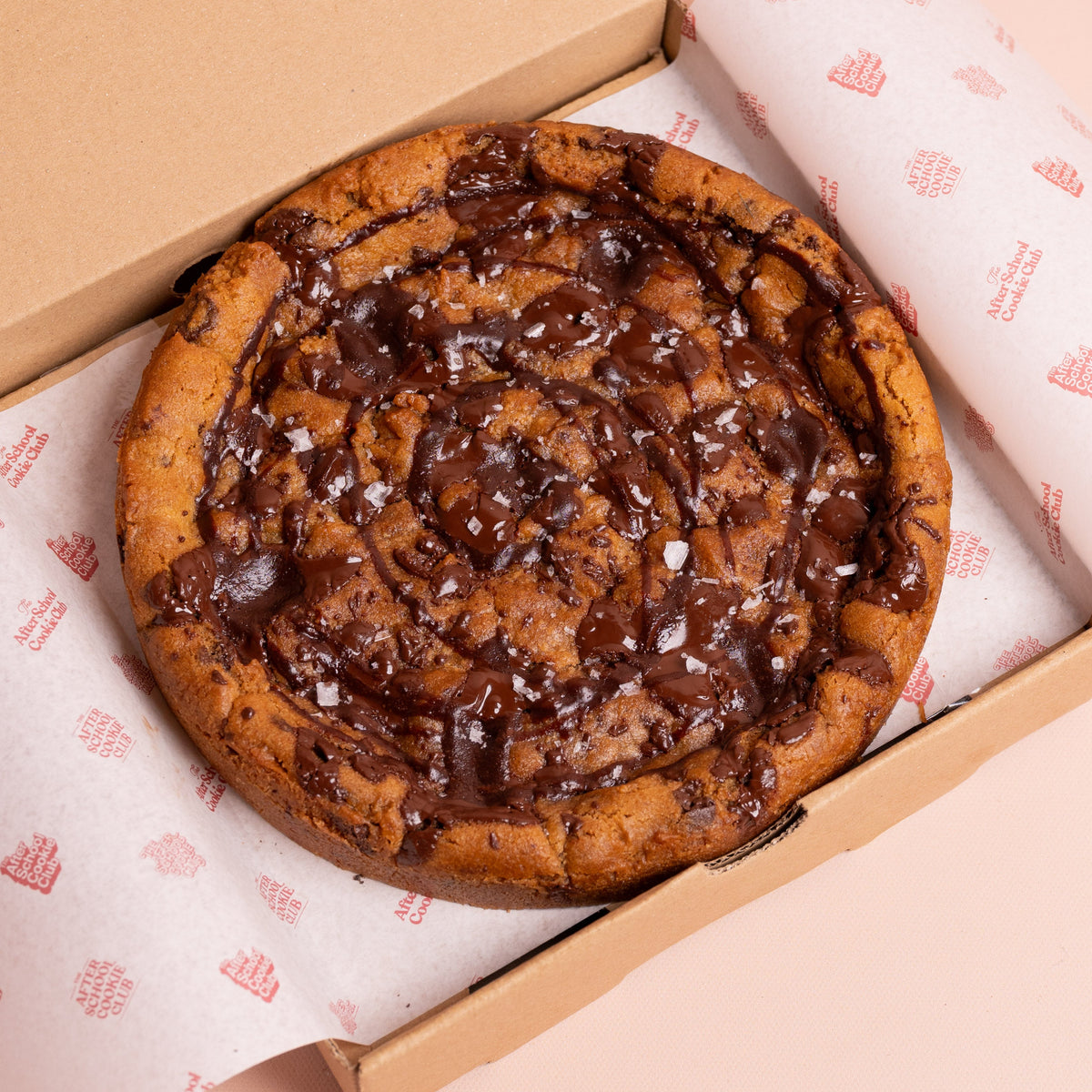 A large chocolate chip cookie with chocolate drizzle and sea salt flakes, presented inside an open brown cardboard box lined with branded white paper.