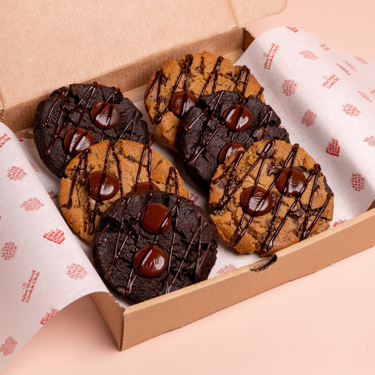 Box of chocolate chip and double chocolate cookies with chocolate drizzle on a pink background