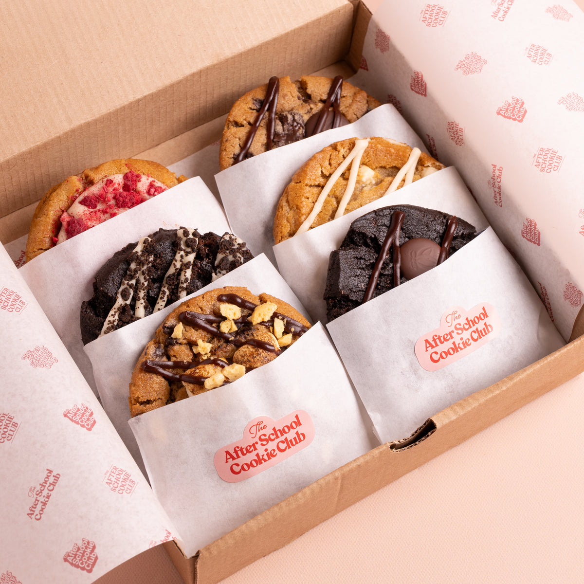 An open cardboard box containing six assorted cookies, each individually wrapped in white paper with a pink logo that reads 'The After-School Cookie Club.' The cookies have varied toppings including chocolate drizzle, nuts, cream, and fruit, and are photographed from above on a light pink background.