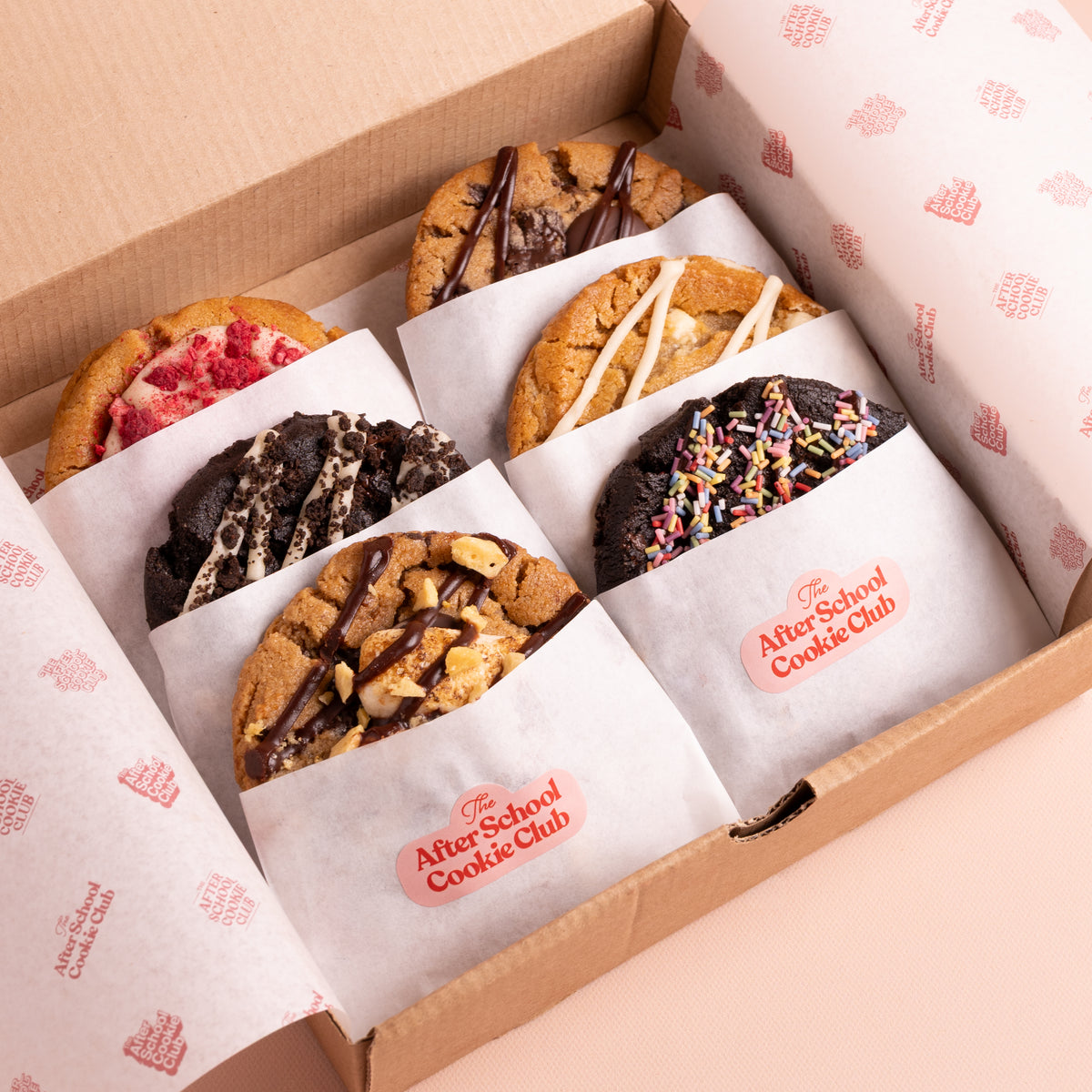 An open cardboard gift box containing six assorted cookies, each individually wrapped in white paper with a pink logo that reads 'After School Cookie Club.' The cookies include chocolate chip, cookie dough, and chocolate-drizzled varieties, with one pink-topped cookie and one sprinkled cookie visible.