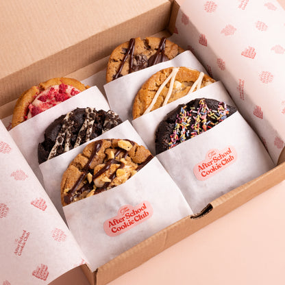 An open cardboard gift box containing six assorted cookies, each individually wrapped in white paper with a pink logo that reads 'After School Cookie Club.' The cookies include chocolate chip, cookie dough, and chocolate-drizzled varieties, with one pink-topped cookie and one sprinkled cookie visible.