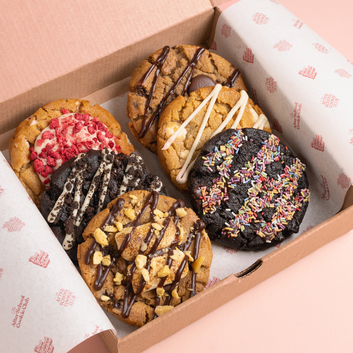 A box of assorted gourmet cookies with various toppings like chocolate drizzle, sprinkles, presented in a cardboard box lined with branded paper.