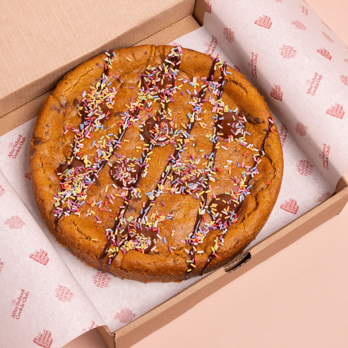 A large chocolate chip cookie with drizzled chocolate sauce and colorful sprinkles, presented inside a brown cardboard box lined with branded white paper.