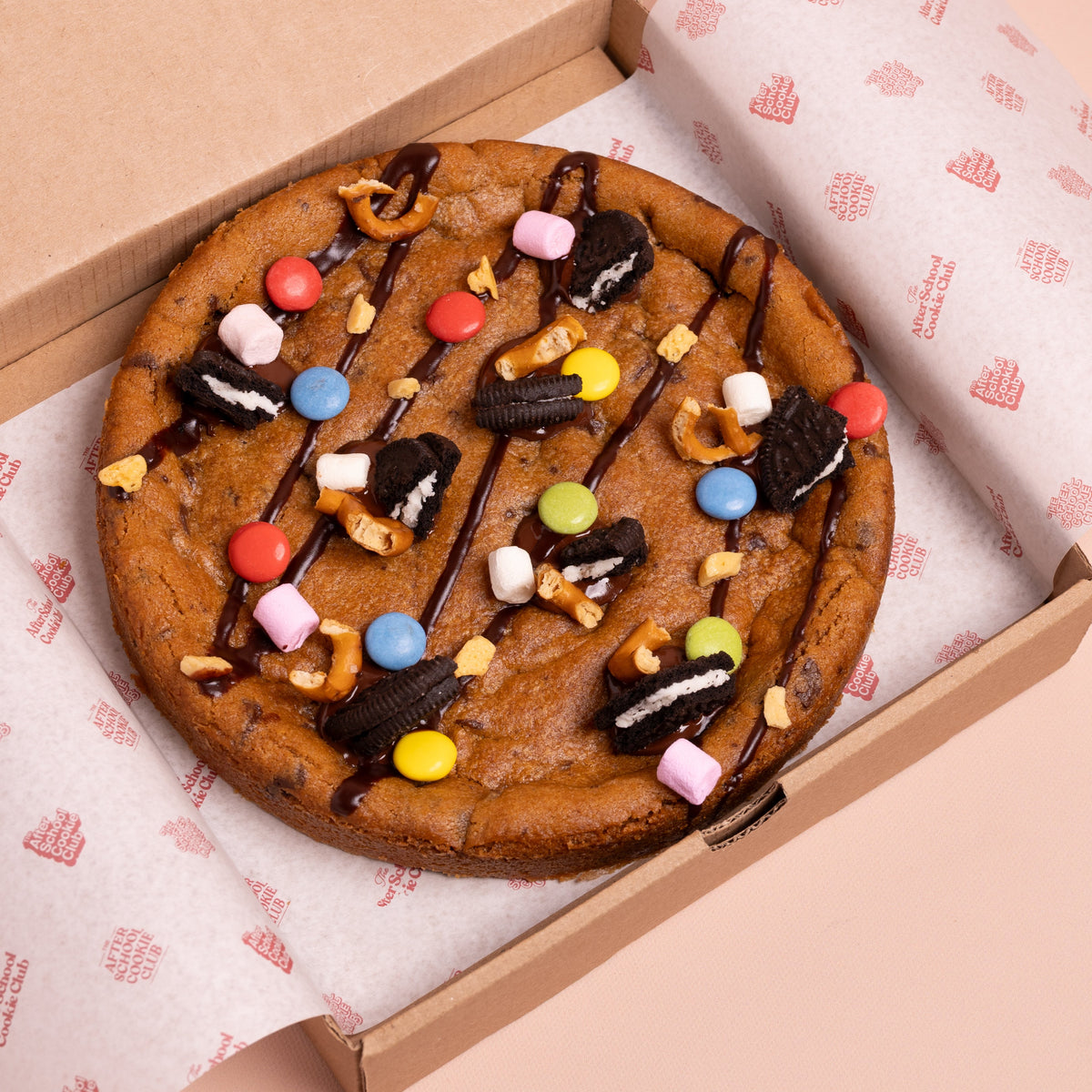 A large chocolate chip cookie  topped with chocolate drizzle, Oreo-style cookies, pretzels, marshmallows, and colourful chocolate buttons, presented in an open cardboard box lined with branded paper.