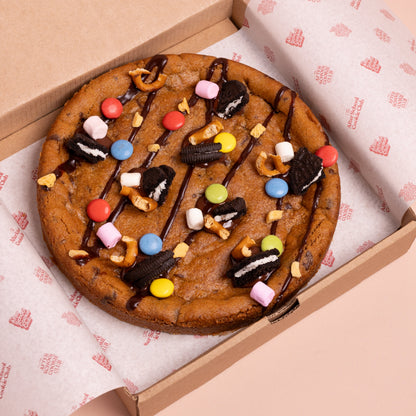 A large chocolate chip cookie  topped with chocolate drizzle, Oreo-style cookies, pretzels, marshmallows, and colourful chocolate buttons, presented in an open cardboard box lined with branded paper.