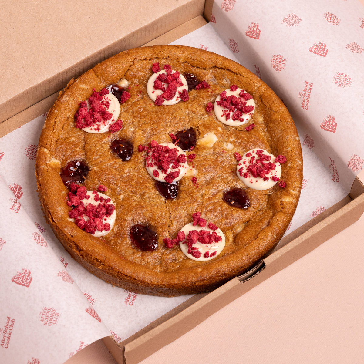 A round chocolate chip cookie with white chocolate and raspberry toppings sits inside an open brown cardboard box lined with branded white paper.