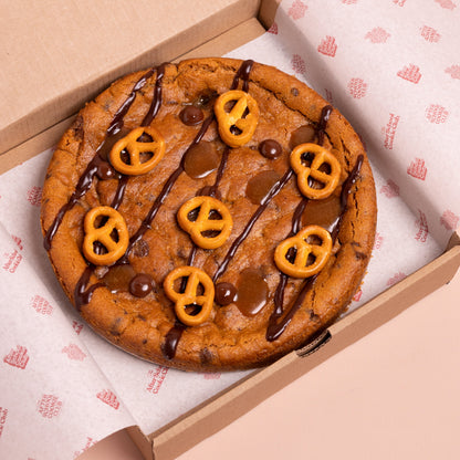 A giant chocolate chip cookie topped with chocolate drizzle, caramel sauce, and mini pretzels, presented inside an open cardboard box lined with branded paper.