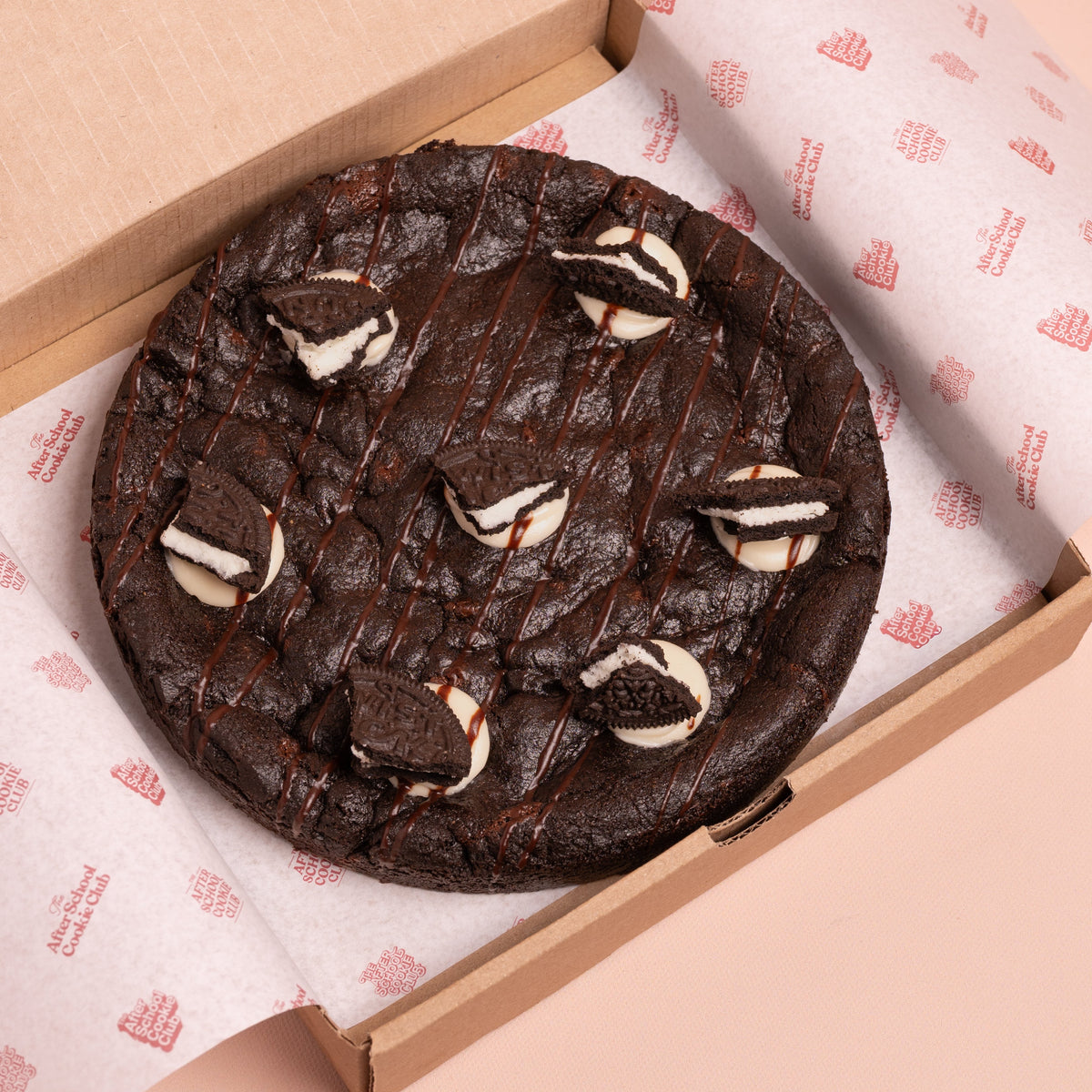 A giant double chocolate cookie with Oreo-style cream and cookie pieces, presented inside a brown cardboard box lined with branded white paper on a pale pink background.