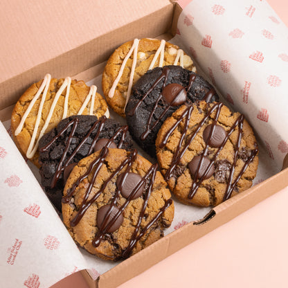 A box of assorted chocolate chip cookies, including chocolate drizzle, white drizzle, and dark chocolate chip varieties, arranged in a brown cardboard box lined with branded paper.