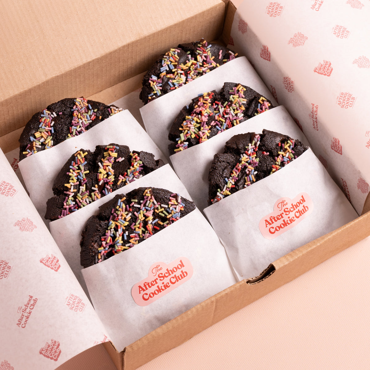 A box of six double chocolate chip cookies topped with colourful sprinkles, individually wrapped in white paper with the 'AfterSchool Cookie Club' logo, presented in a brown cardboard box.