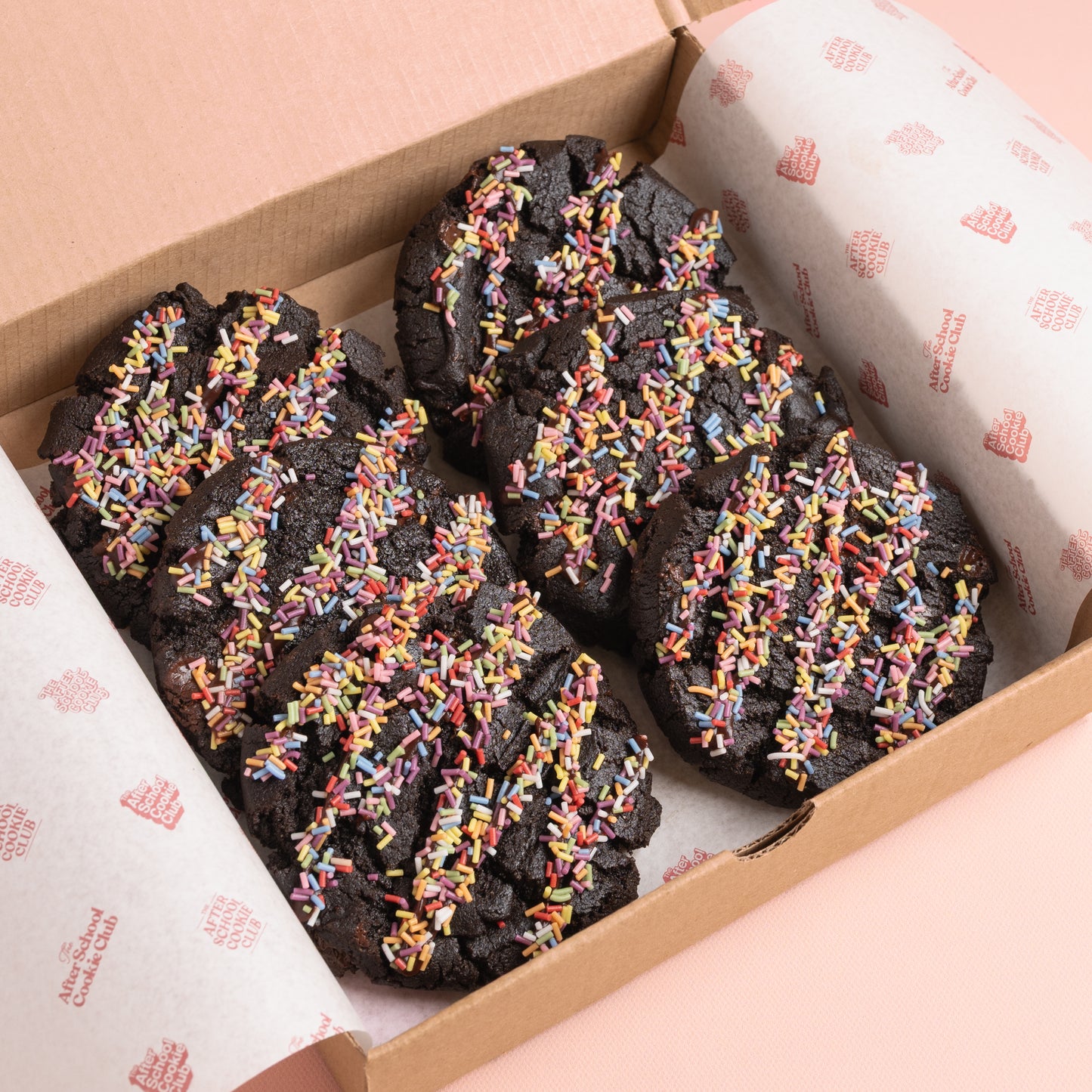 Four double chocolate chip cookies topped with colourful sprinkles, presented in a brown cardboard box lined with branded white paper.