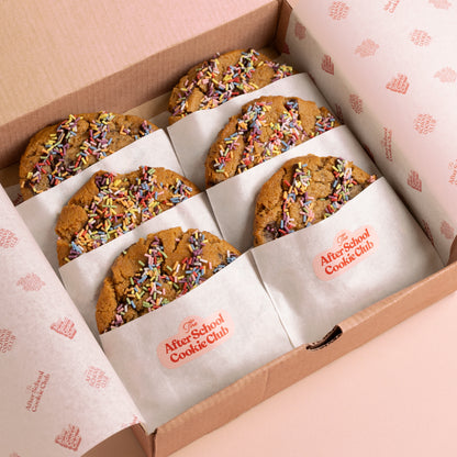 An open cardboard box of six chocolate chip cookies topped with colorful sprinkles, each wrapped in branded After School Cookie Club parchment paper.