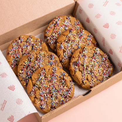 Six chocolate chip cookies topped with colorful sprinkles, arranged inside a brown cardboard gift box lined with branded white paper.