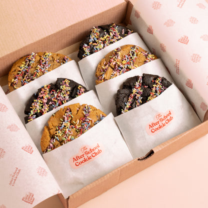 Six assorted cookies in a cardboard gift box lined with branded paper. The cookies are arranged in two rows of three, with three double chocolate chip cookies and three choc chip cookie, each topped with colourful rainbow sprinkles.