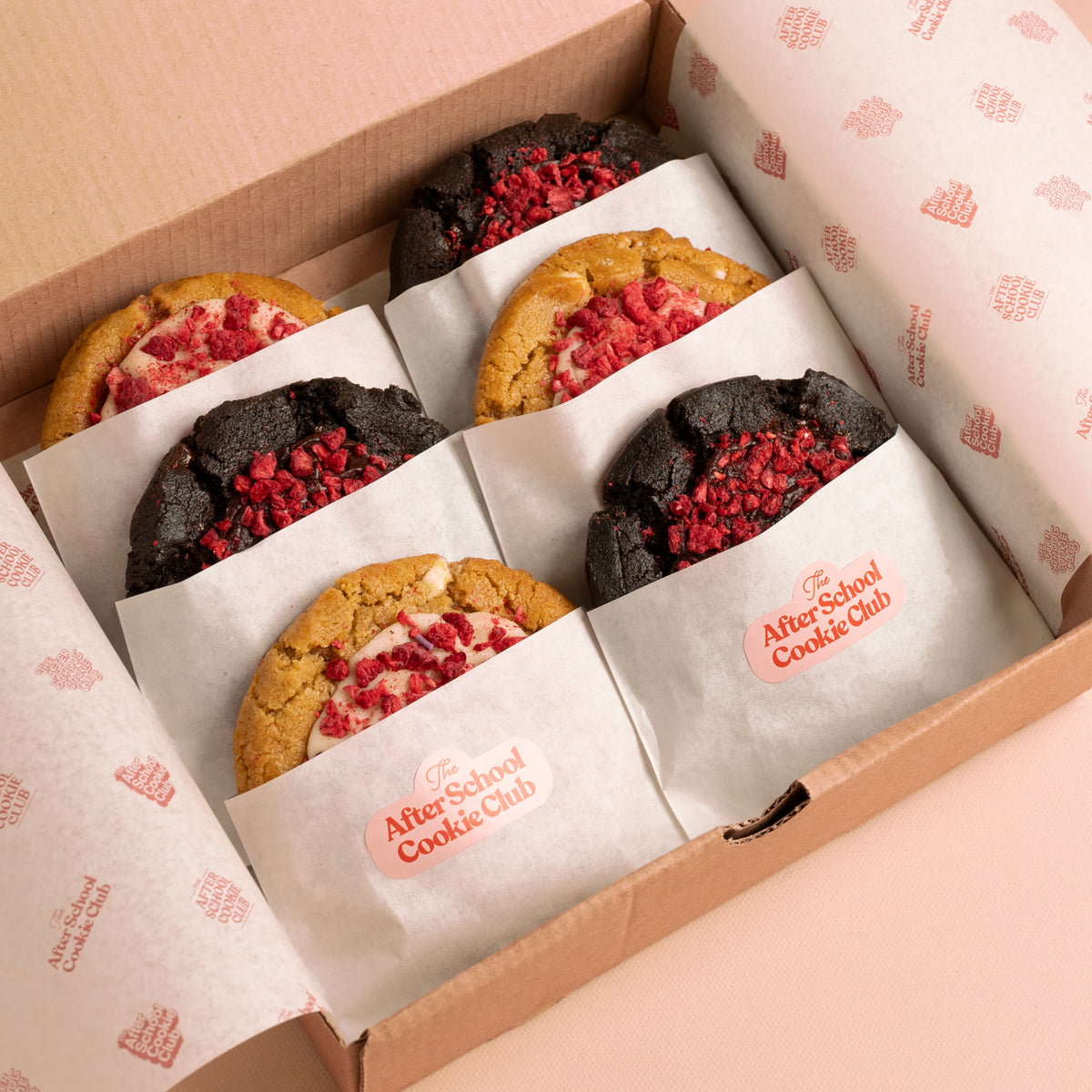 An open cardboard gift box lined with branded After School Cookie Club tissue paper, containing six assorted cookies wrapped in white paper sleeves. Three cookies are double chocolate chip style with red raspberry topping, and three are white chocolate cookies with white chocolate ganache and red raspberry topping.