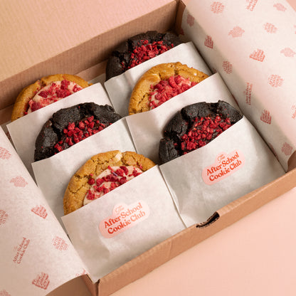 An open cardboard gift box lined with branded After School Cookie Club tissue paper, containing six assorted cookies wrapped in white paper sleeves. Three cookies are double chocolate chip style with red raspberry topping, and three are white chocolate cookies with white chocolate ganache and red raspberry topping.