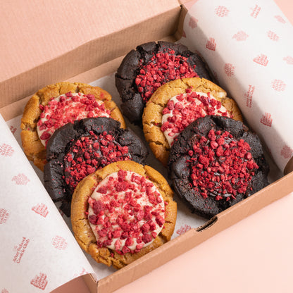 An open cardboard gift box lined with branded After School Cookie Club tissue paper, containing six assorted cookies. Three cookies are double chocolate chip style with red raspberry topping, and three are white chocolate cookies with white chocolate ganache and red raspberry topping.